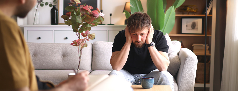 Ein Mann mit erschöpftem Gesichtsausdruck sitzt auf einem Sofa und stützt seinen Kopf in seinen Händen ab.